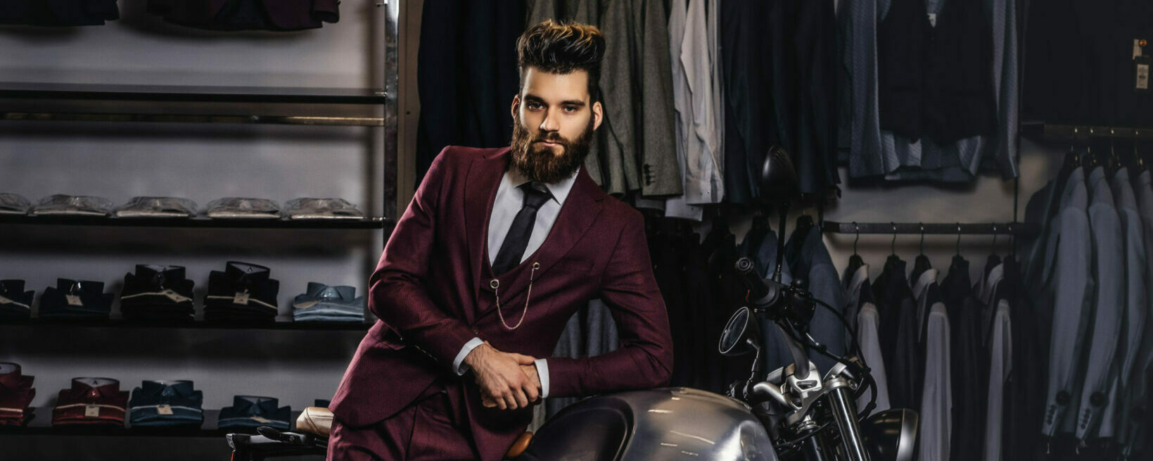 Elegantly dressed handsome man with stylish beard and hair leaning on retro sports motorbike at men's clothing store.